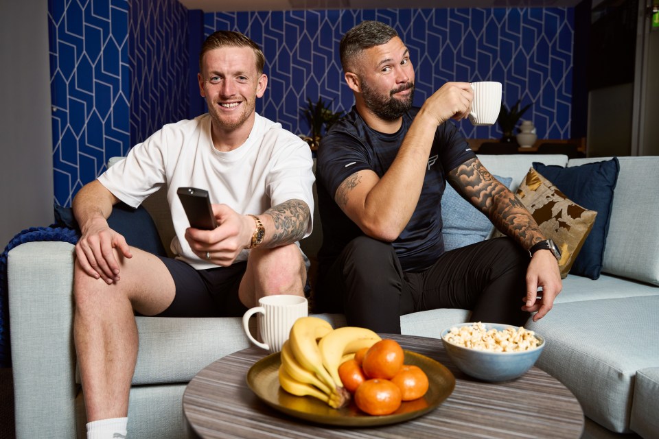 two men sit on a couch drinking coffee and eating fruit