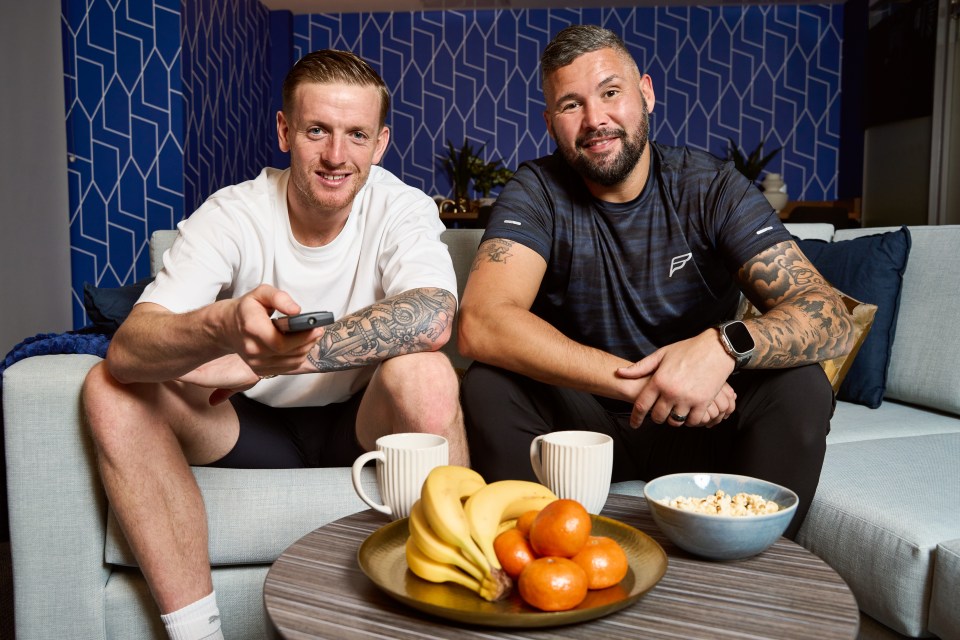 two men sit on a couch with a plate of fruit and popcorn