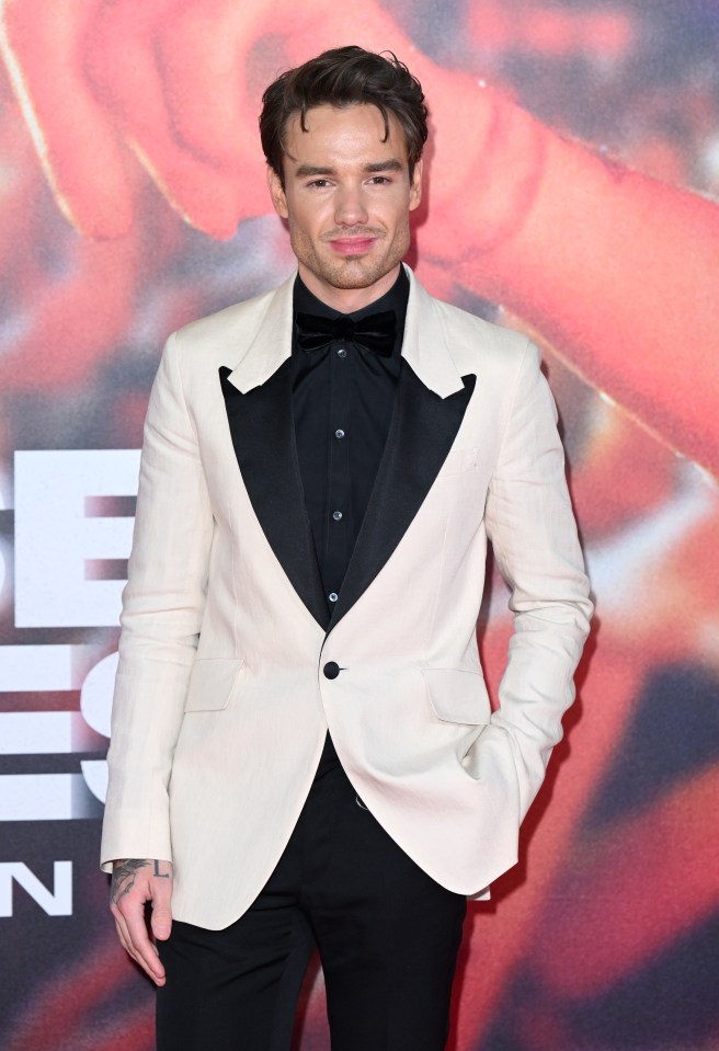 a man in a white suit and black bow tie stands on a red carpet