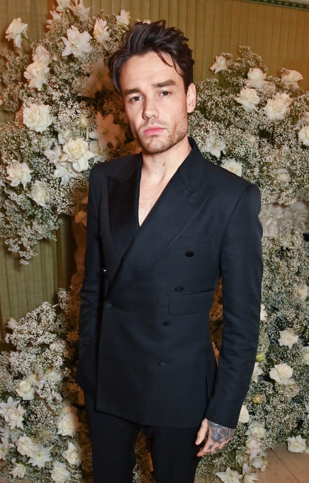 a man in a black suit stands in front of a wall of white flowers