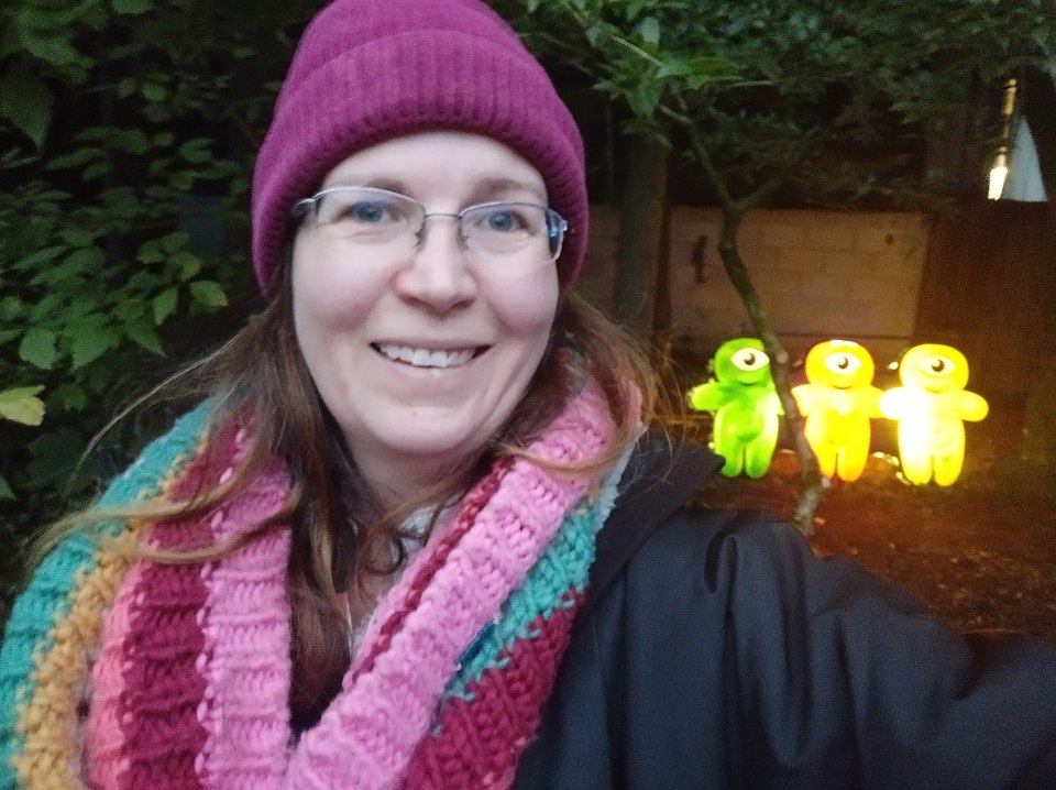 a woman wearing a purple beanie and a pink scarf smiles while standing in front of glowing stuffed animals