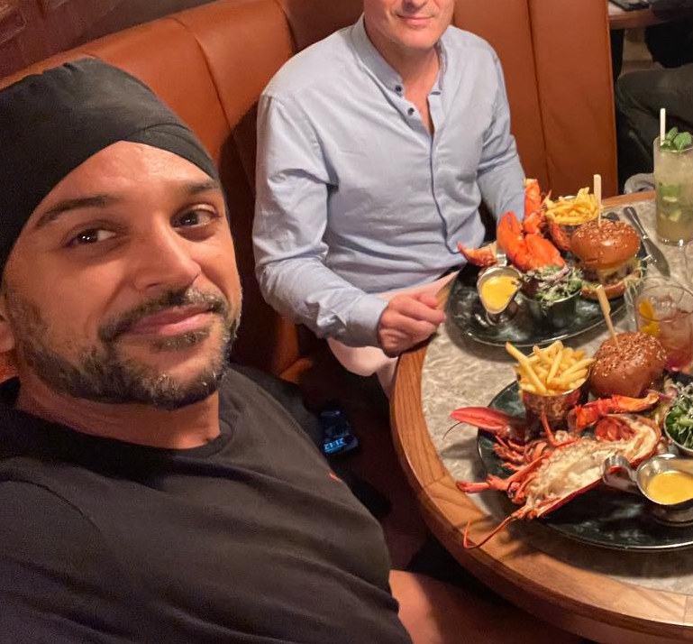 two men are sitting at a table with plates of food including lobsters and hamburgers