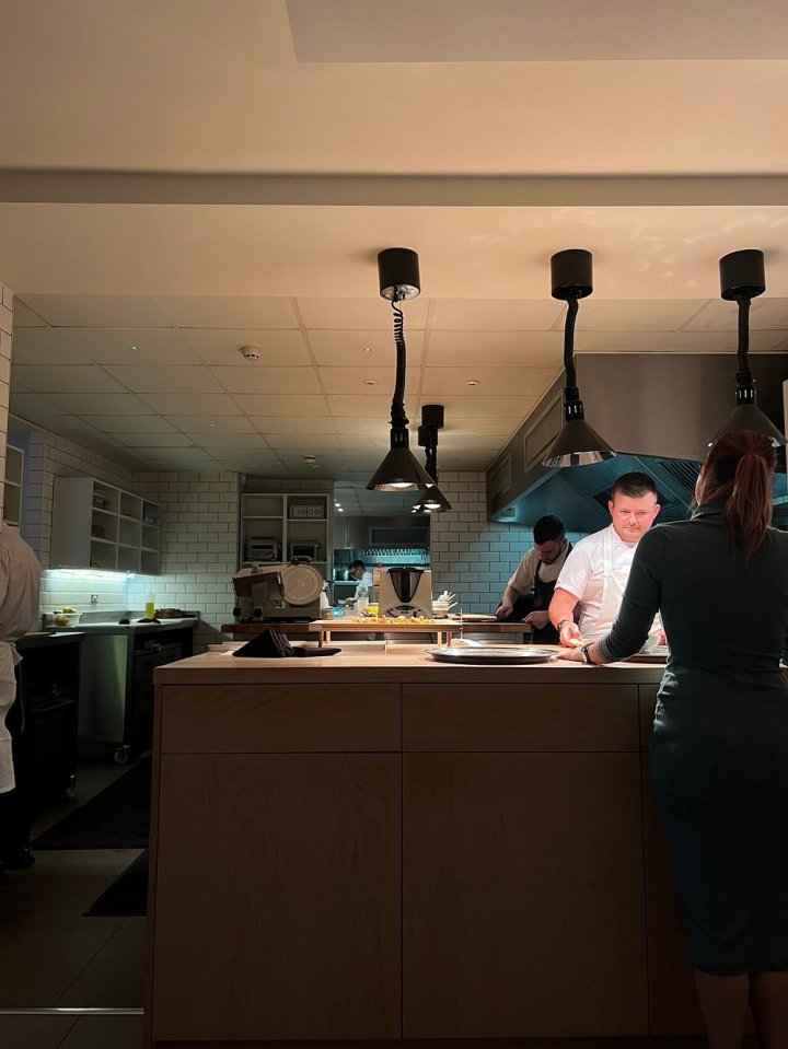 a woman stands in a kitchen with chefs preparing food