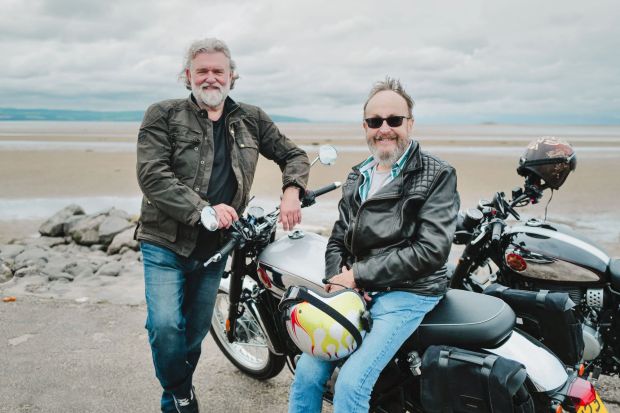 two men standing next to motorcycles with one wearing a helmet with flames on it