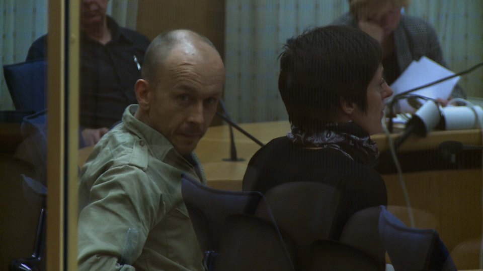 a man and a woman sit in a courtroom behind a glass wall