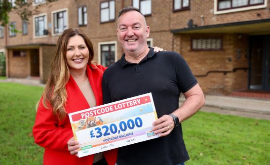 a man and a woman are holding a postcode lottery cheque in front of a house .