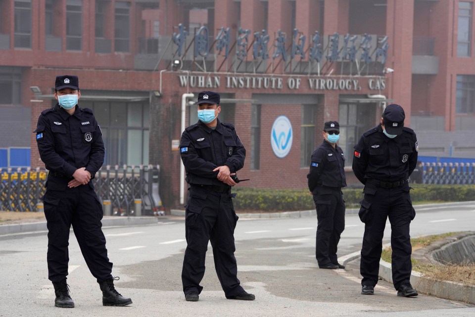 four police officers wearing masks stand in front of a building that says wuhan institute of virology