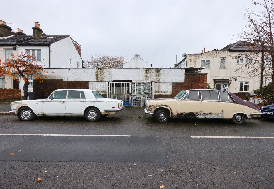 A white old car is parked next to a brown rusty car nearby in front of an abandoned garage