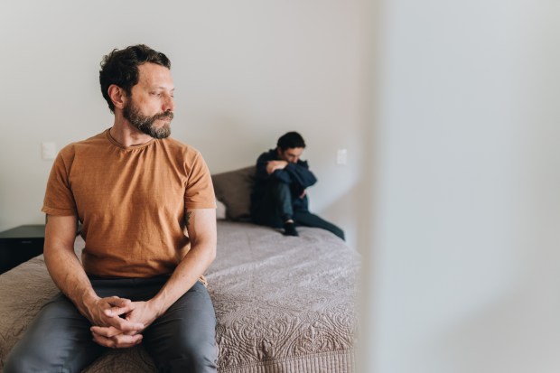 a man in a brown shirt sits on a bed next to another man