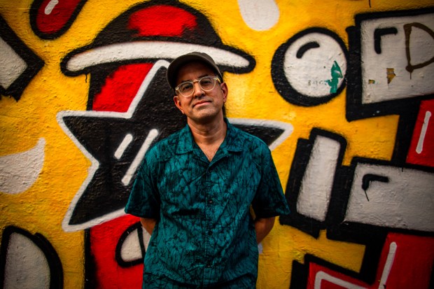 a man wearing glasses and a hat stands in front of a wall with graffiti on it