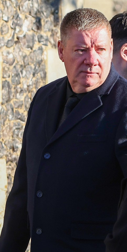 a man in a black coat and tie stands in front of a stone wall