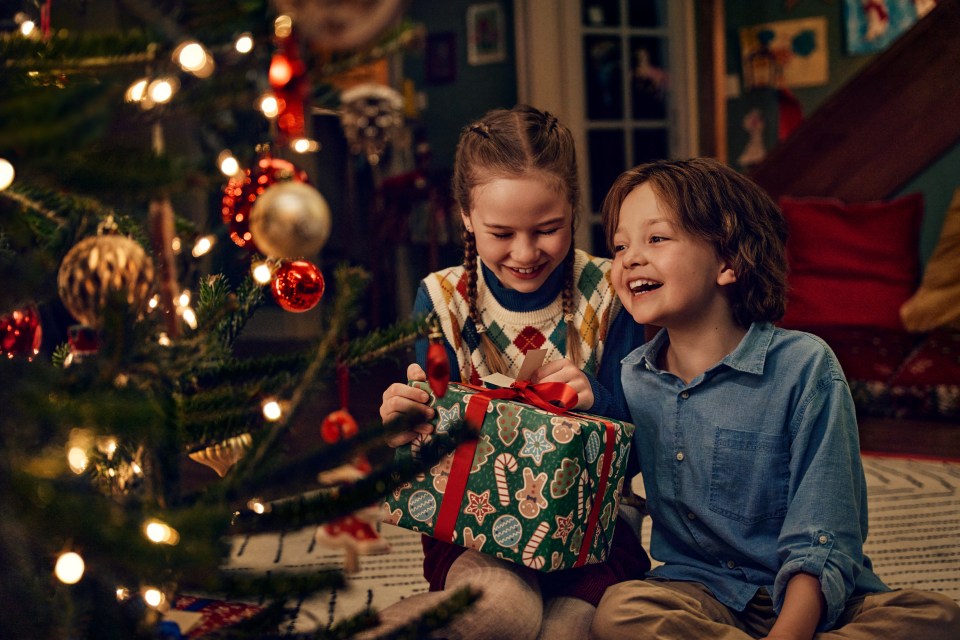 a boy and a girl are sitting in front of a christmas tree opening presents