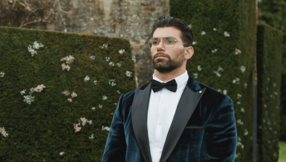 a man in a tuxedo and bow tie stands in front of a hedge