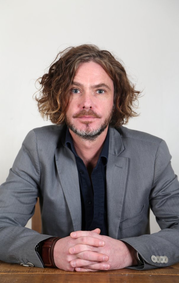 a man with curly hair and a beard sits at a table with his hands folded