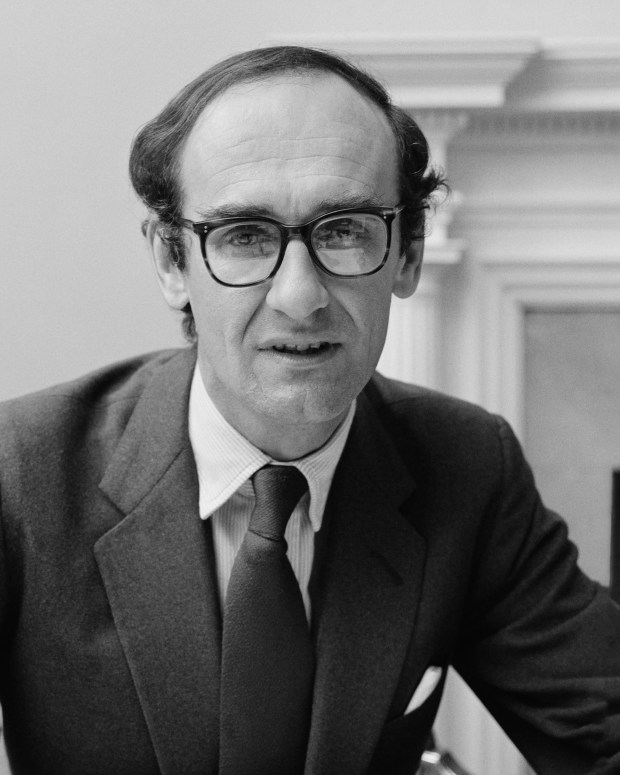 a man wearing glasses and a suit stands in front of a fireplace