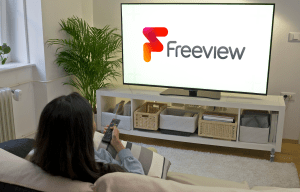 a woman sits on a couch watching a tv with the freeview logo on the screen