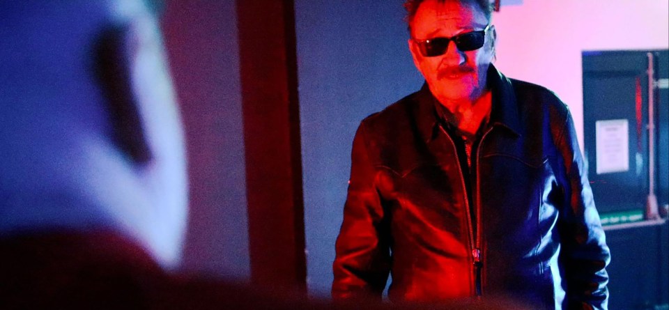 a man wearing sunglasses and a leather jacket stands in a dark room