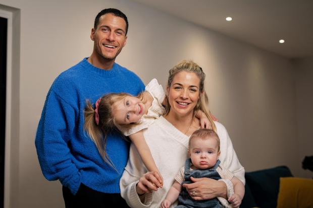 a man in a blue sweater stands next to a woman holding a baby