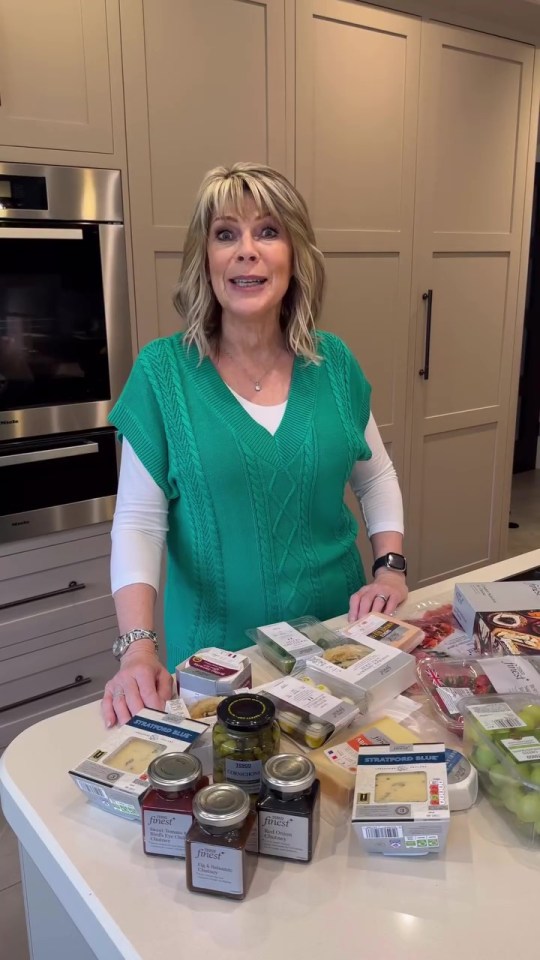 a woman in a green sweater is standing in front of a counter full of food including jars of jam