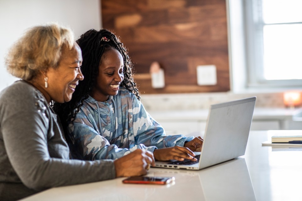 two women sit at a table looking at a laptop