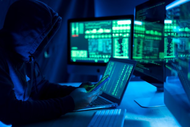 Hooded figure using a laptop and phone, surrounded by computer screens displaying code.