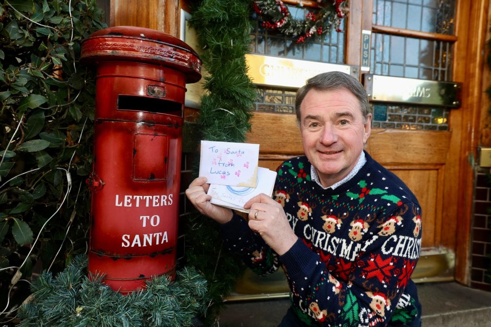 Man in Christmas sweater holding letters to Santa by a Santa letterbox.