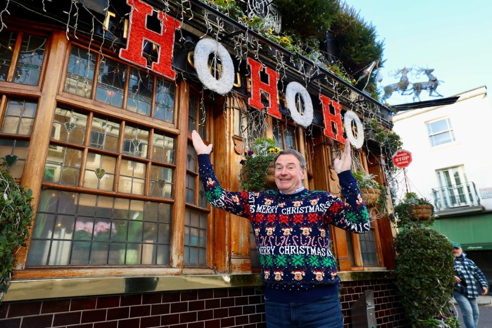 Man in Christmas sweater stands in front of a pub decorated with "Ho Ho Ho" sign.