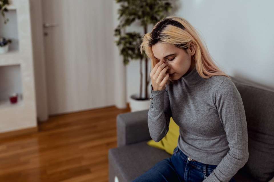 Poor young Caucasian woman sitting on sofa touching her nose having sinus problems