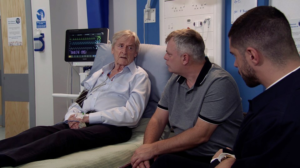 an elderly man laying in a hospital bed talking to two men