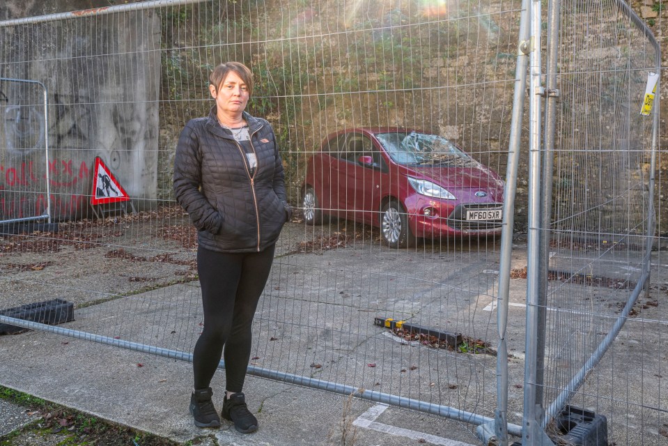 a woman stands in front of a fence with a red car parked behind it