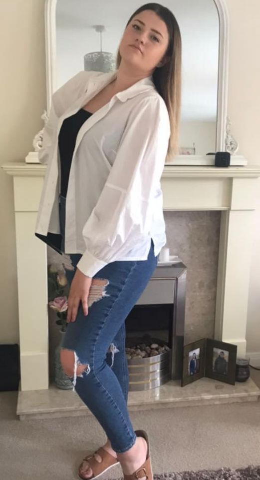 a woman in a white shirt and ripped jeans stands in front of a fireplace