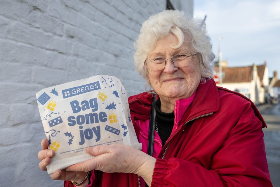 A senior woman smiling while holding a Greggs bag.