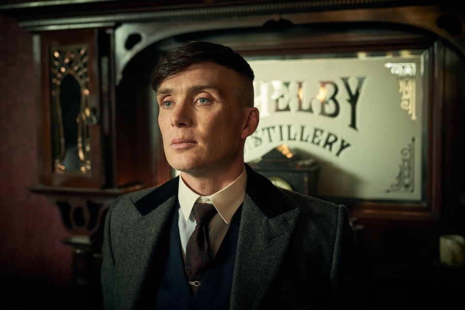 A photo of Tommy Shelby (Cillian Murphy) standing in a dark room looking at a stillery. He is wearing a dark suit and a cap. The photo is taken from the side.