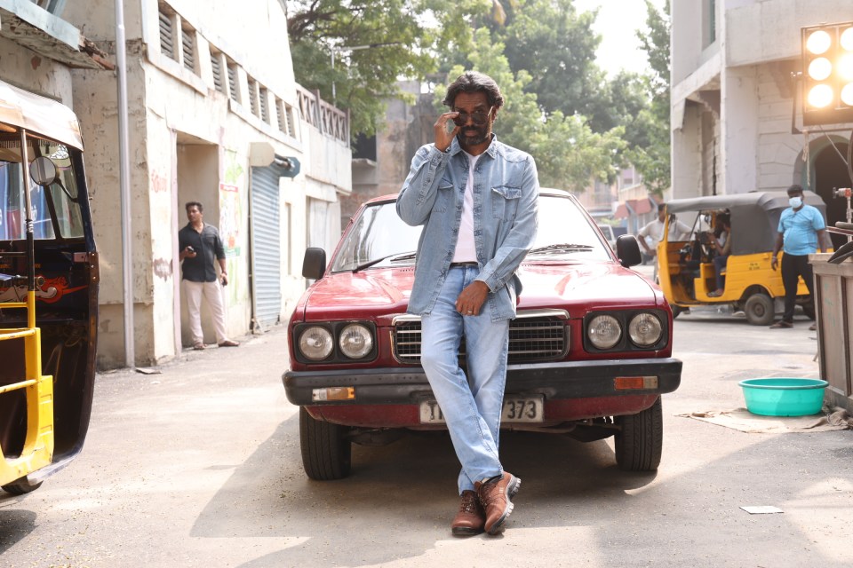 Man in denim jacket leans against a vintage car in a city street.