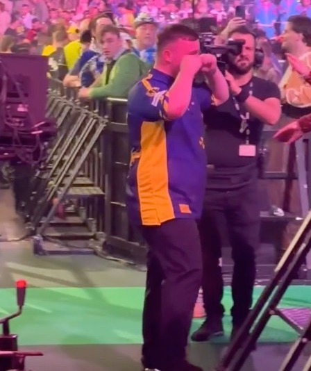 A man in a purple and gold jersey shields his eyes while standing near a cameraman in a large crowd.