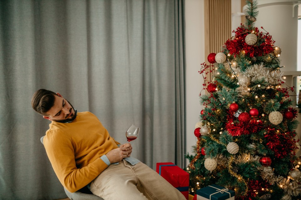 Drunk man sleeping on the chair next to the Christmas tree with Christmas gifts and a wine glass in his hands. Tired guy after a Christmas party concept.