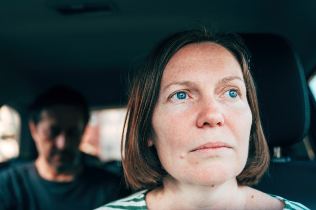 Close-up of a dissatisfied woman in a car, with a blurred man in the background.