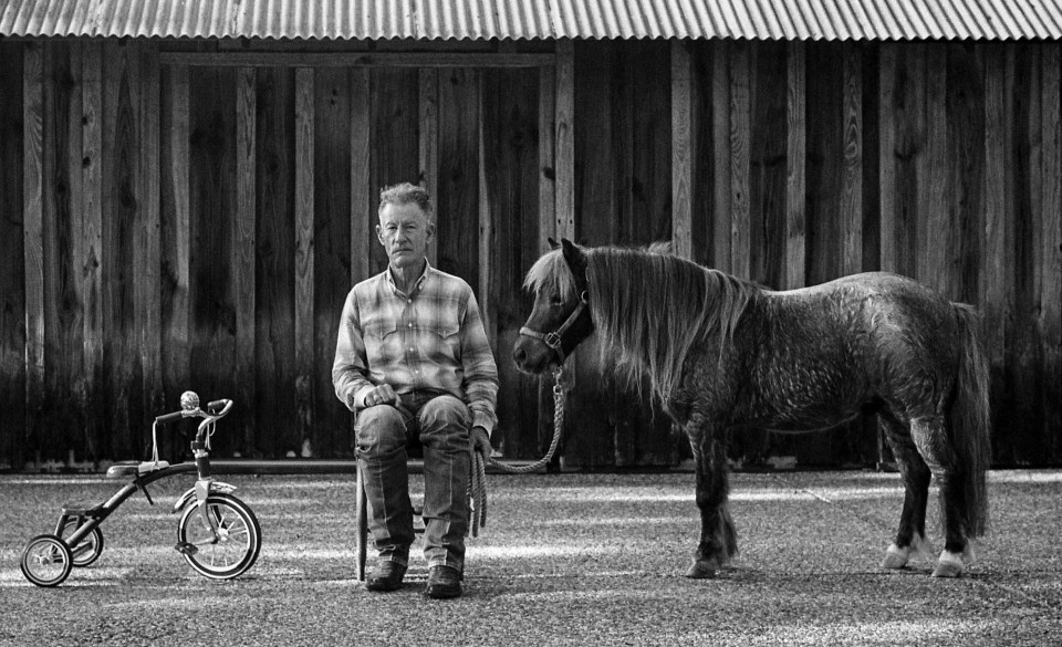 Black and white photo of Lyle Lovett sitting in front of a wooden barn with a pony.  A tricycle is next to him.