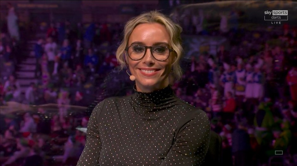 A woman wearing glasses presents live darts coverage.