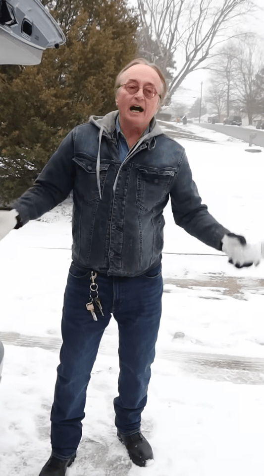 A man in a denim jacket stands in the snow, gesturing toward the open trunk of a car.