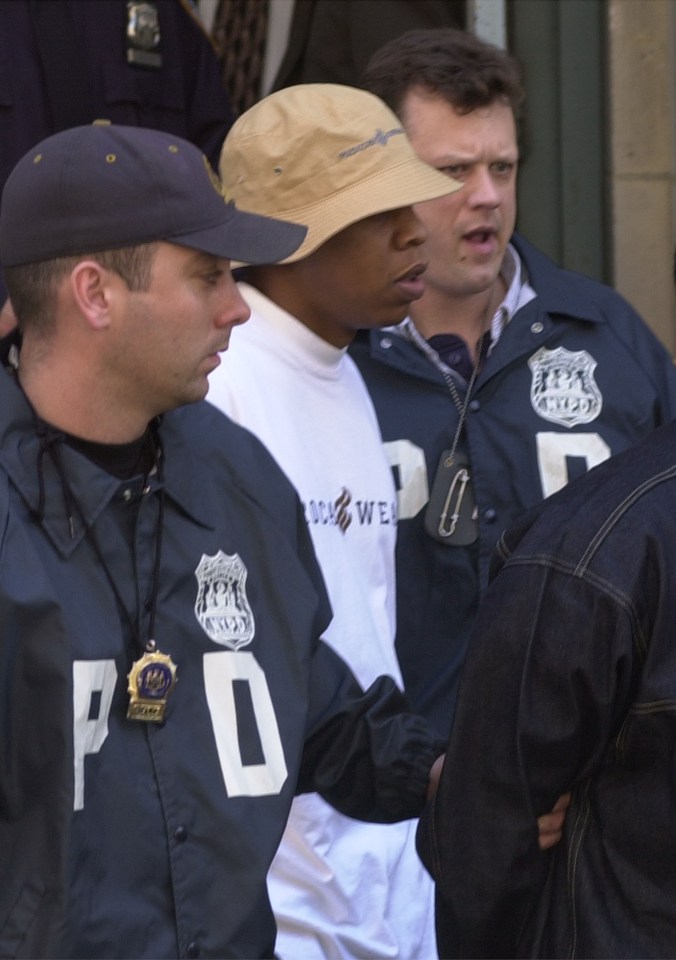 A man wearing a white shirt, black pants, and a black hat is being led out of a precinct station by police in New York City.