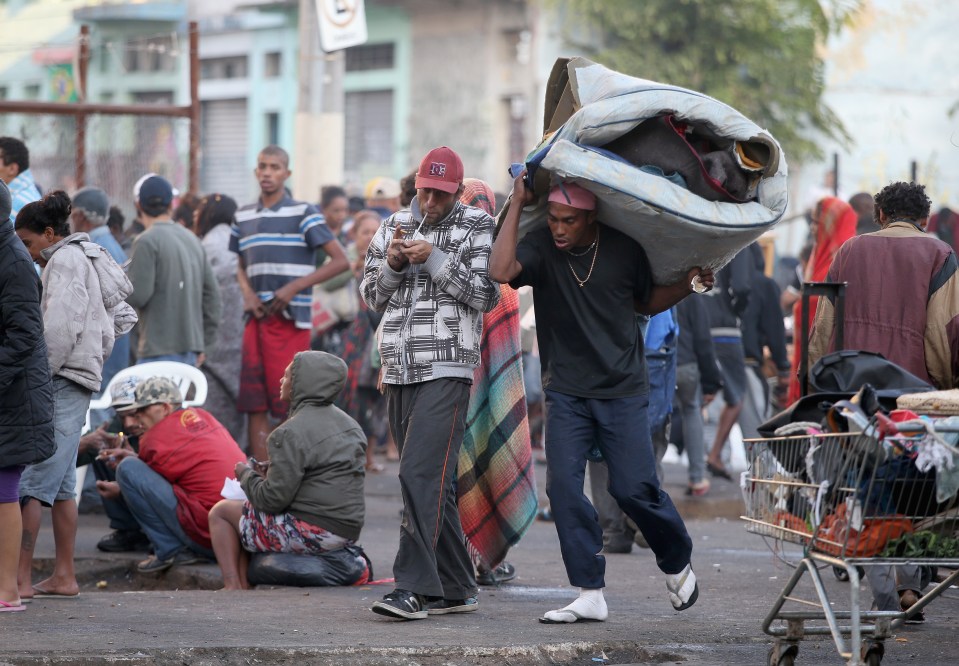 A man stands with arms outstretched in Cracolandia, a drug-ridden area of Sao Paulo, Brazil.  Surrounding him are people and belongings in a scene of poverty and urban decay.