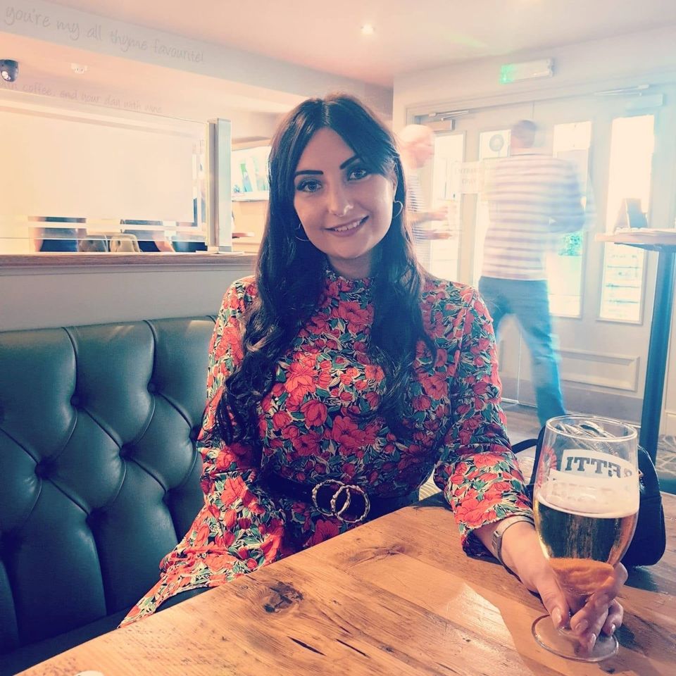 Woman in floral dress sits at a table in a pub, holding a beer.