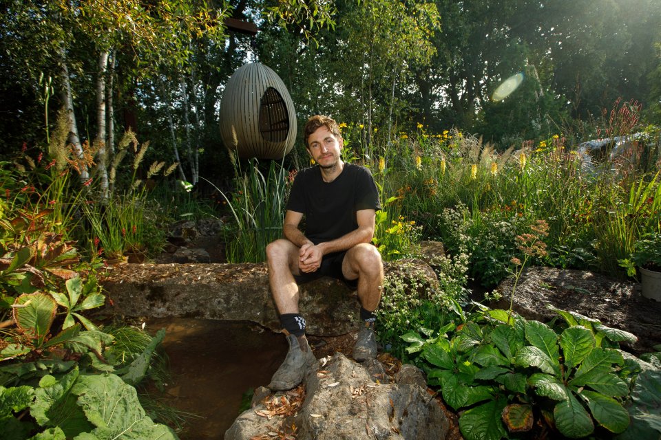 A garden designer sits on a rock in a garden he designed.