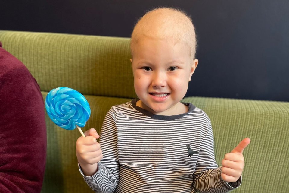 Smiling boy with shaved head holds a blue lollipop and gives a thumbs up.