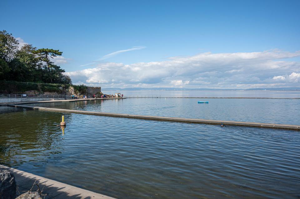 Outdoor saltwater swimming pool with people swimming and a town in the background.