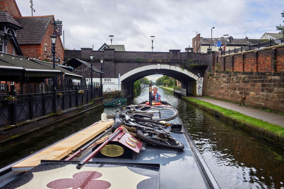 Woman steering a narrowboat under a bridge.