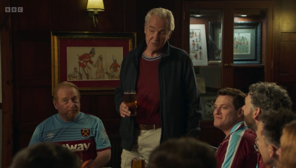 A group of men, some wearing West Ham United jerseys, sit at a table in a pub; one man stands, holding a pint of beer.