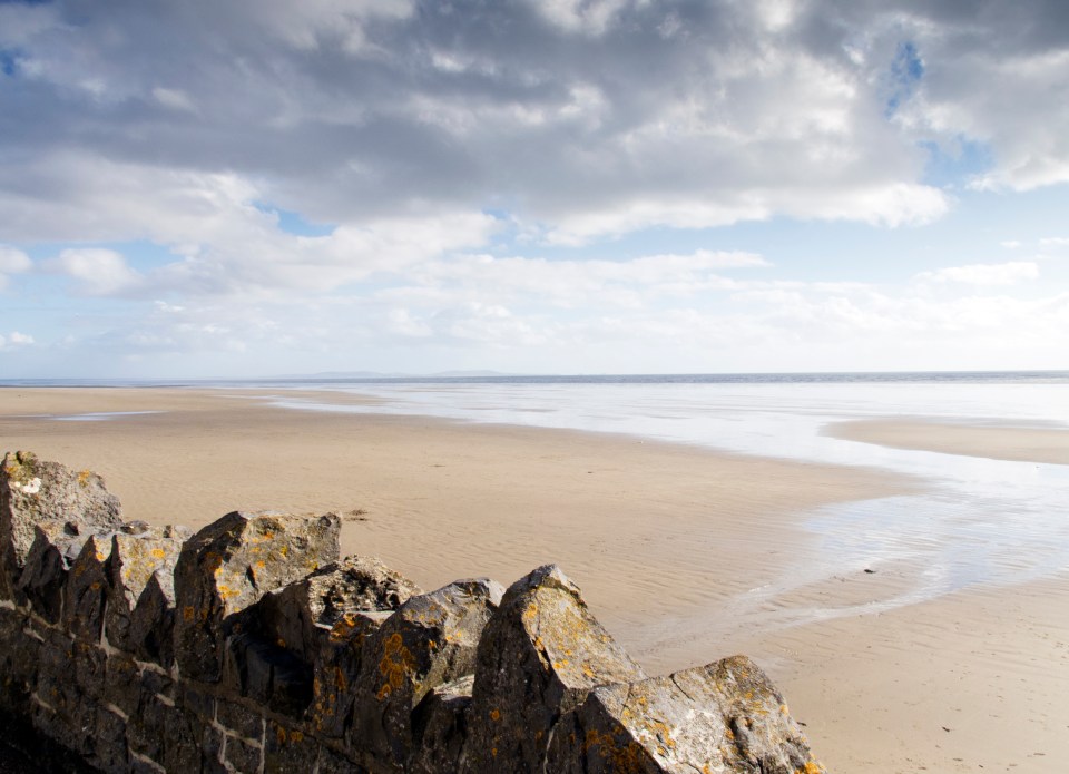 Stone wall overlooking a sandy beach and calm sea under a partly cloudy sky.