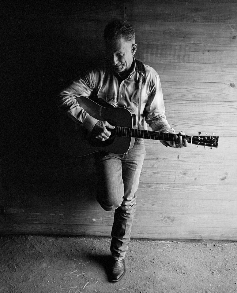 Black and white photo of Lyle Lovett playing an acoustic guitar.